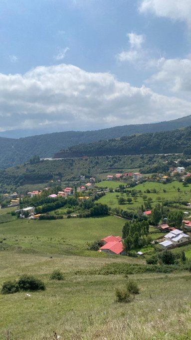 A scenic landscape view of rolling green hills, scattered houses with red roofs, and forested mountains under a partly cloudy blue sky.