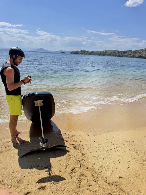 A man wearing a helmet and yellow shorts stands on a sandy beach holding a controller, with a hydrofoil board and paddle in the sand nearby, with mountains and ocean in the background.