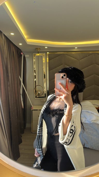 A mirror selfie of a woman wearing a black fuzzy hat, black top, and white knit cardigan in a bedroom setting.