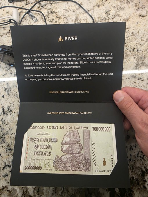 A hand holds open a black presentation card from 'RIVER' featuring a Zimbabwean hyperinflated 200 million dollar banknote, with text explaining how the banknote illustrates why Bitcoin was designed with a fixed supply.