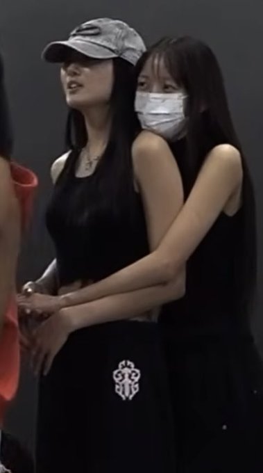 Two young women pose together, one wearing a gray baseball cap and black crop top, the other with long dark hair, a white face mask, and a black tank top featuring a white crest design.