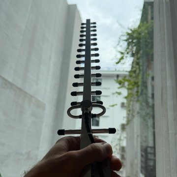 A first-person view of a hand holding a dark, comb-like metal tool outdoors, with white curtains and a building visible in the background.