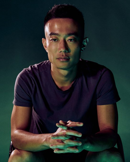 A studio portrait of a young man with short dark hair, wearing a dark t-shirt, sitting with his hands clasped against a teal and dark green background.