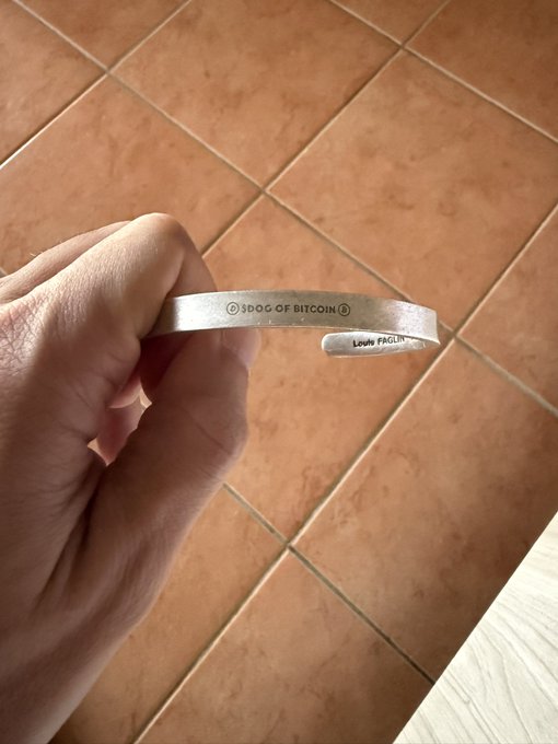 A close-up of a hand holding a silver cuff bracelet engraved with '② SPOOG OF BITCOIN ②' and 'Louis FAGLIN' against a terracotta tile floor.