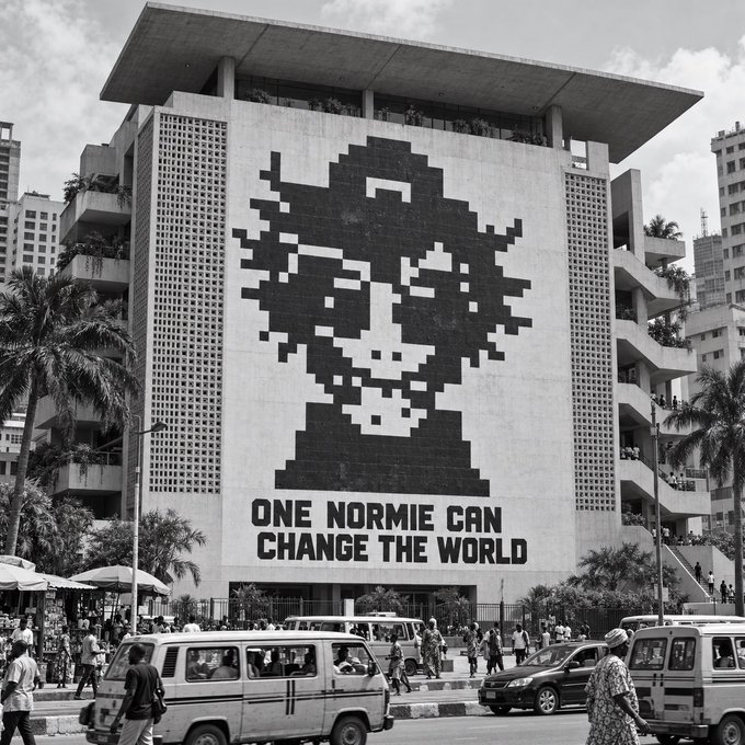 A black and white photograph of a large building with a pixelated mural of a person's face and the text "ONE NORMIE CAN CHANGE THE WORLD" on the front, with cars and pedestrians in the foreground.