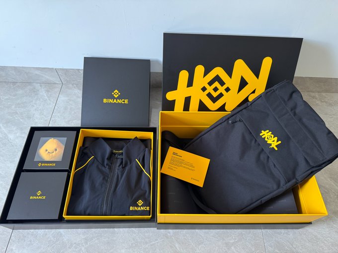 Two unboxed merchandise sets featuring a black Binance jacket and a black HODL backpack in black and yellow packaging.