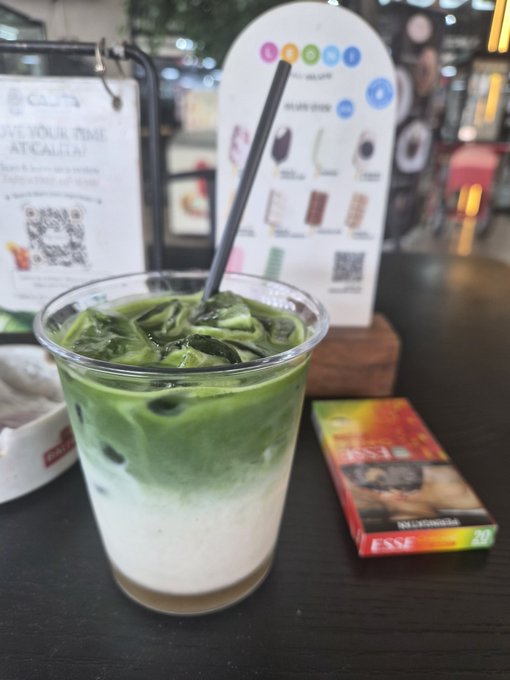 A vibrant green matcha latte in a clear plastic cup with a black straw sits on a dark table next to an "ESSE" cigarette pack, with a "GIVE YOUR TIME AT CALIA" sign featuring a QR code visible in the background. The casual cafe setting complements the post's message about taking it easy and using the OKX virtual card for simple, hassle-free snack purchases.