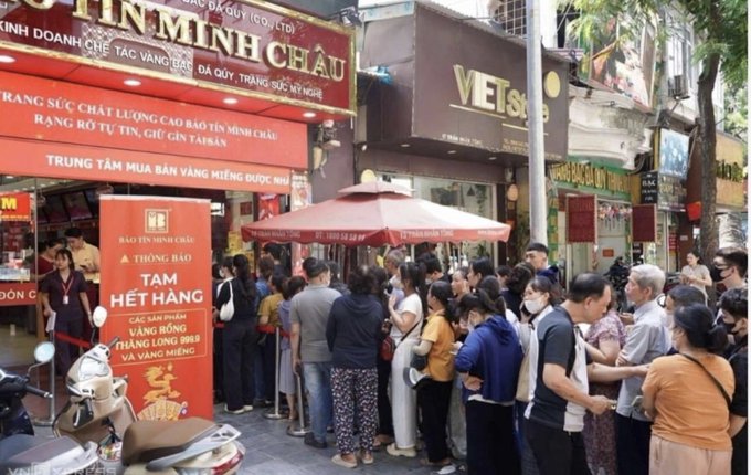 A large crowd of people queues outside a Vietnamese gold shop, with banners advertising 'Bảo Tín Minh Châu' and 'Trung Tâm Mua Bán Vàng Miếng Được Nh', waiting to purchase gold amidst high demand.
