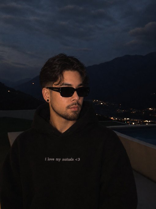 A nighttime portrait of a young man wearing dark sunglasses and a black hoodie that reads 'i love my mutuals <3', with mountains and city lights behind him.