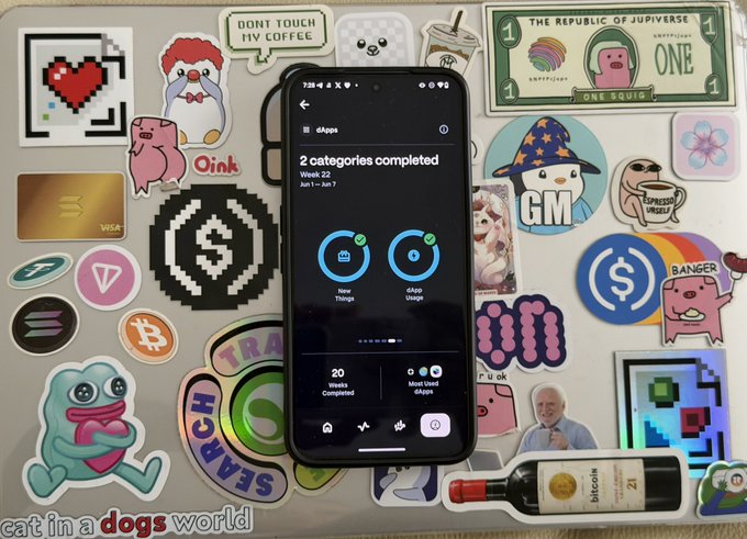 The image features a cell phone with various stickers and logos surrounding it. The phone displays two different categories of completed tasks, including "2 Categories Completed" and "2 Categories Started". There are also other apps visible on the screen, such as "@solanamobile", which is an app for tracking daily steps or physical activity. Additionally, there's a bottle of wine placed next to the cell phone, suggesting that someone might be enjoying their time while using the device.