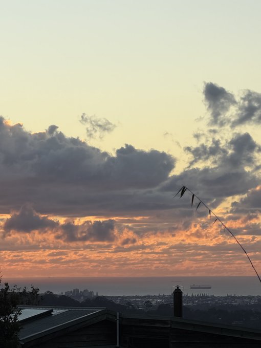 A sunset or sunrise photograph showing pink and orange-hued clouds over a cityscape and ocean, with a roof in the foreground and what appears to be a kite string extending across the sky.