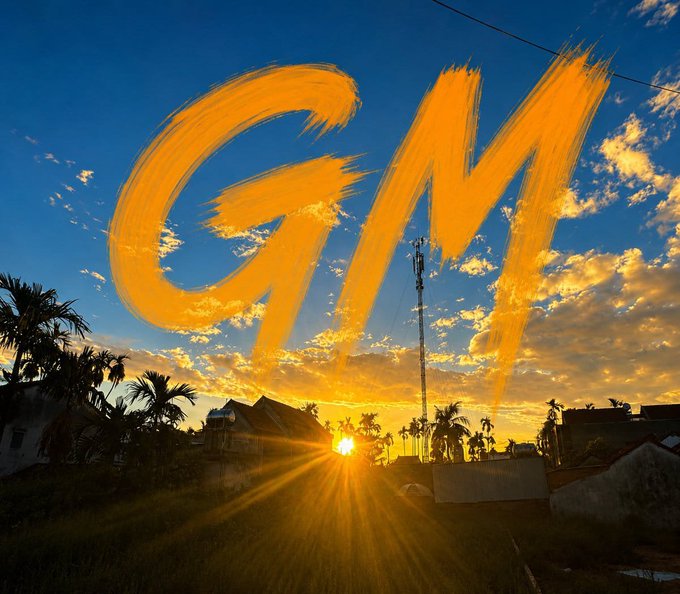 A vibrant sunset photo over a tropical landscape with palm trees and a communications tower, overlaid with a large, brush-stroked orange 'GM' (Good Morning).