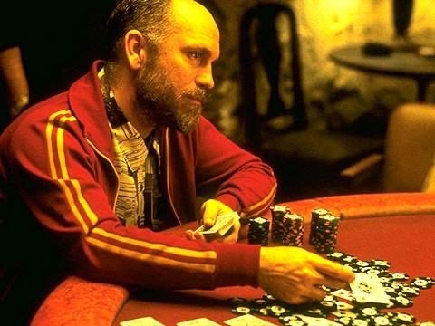 A man in a red track jacket sits at a poker table covered in chips and playing cards, holding his cards thoughtfully.