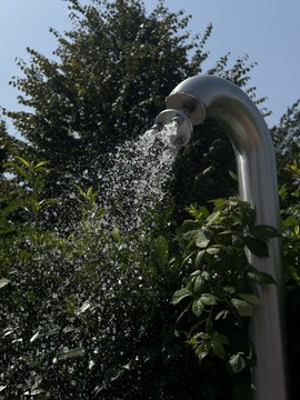 A metal outdoor shower head sprays water downward against a backdrop of lush green trees and a clear blue sky.
