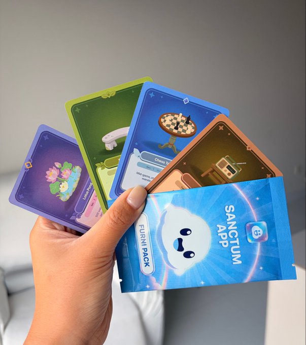 A hand holding several colorful digital collectible cards, including a prominent blue 'Sanctum App' Furni Pack card featuring a cute white bear character, along with green, orange, and purple game cards.