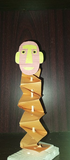 A wooden art sculpture of a stylized figure with a pink and green face and an accordion-folded zigzag body, mounted on a stone base against a dark wood background.