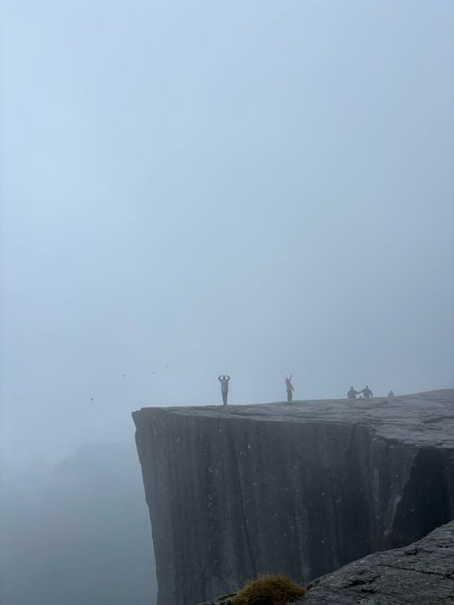 A misty cliff edge scene with small silhouetted figures standing on the rock outcrop against a foggy gray sky.