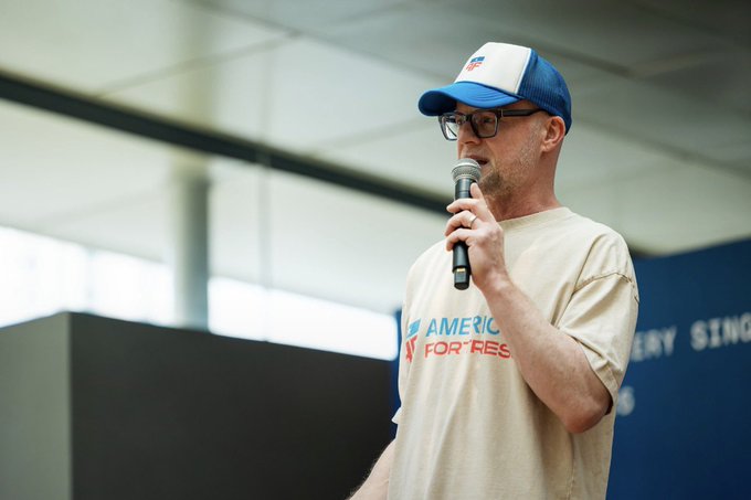 A man wearing glasses, a blue and white cap, and an "America for Rest" t-shirt speaking into a microphone.
