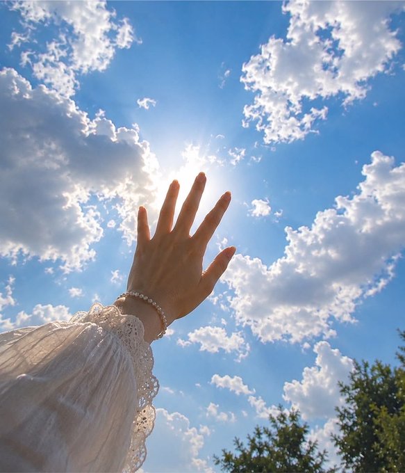 A hand reaches up toward the bright sun against a blue sky filled with scattered clouds.