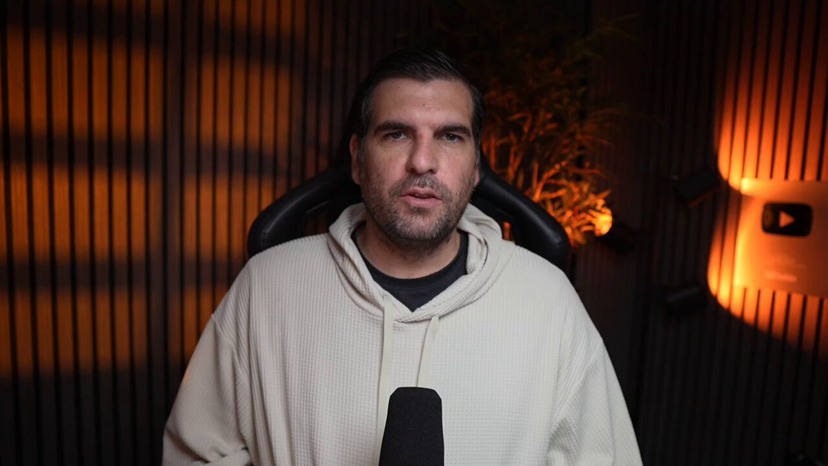 The image shows a man wearing a white hoodie, sitting behind a microphone with his hands on his knees. The background features a dark wall with orange lights, creating an interesting contrast between light and shadow. There are also some speakers visible in the scene. The man appears to be speaking into the microphone or holding it up to someone's ear, possibly discussing the real utility of Ripple Coin or sharing insights about its future adoption.