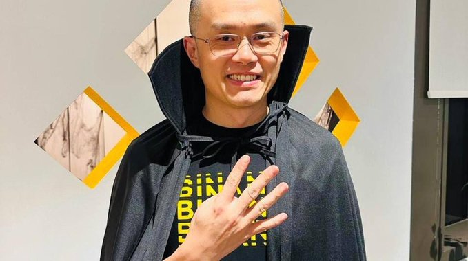 Changpeng Zhao, also known as "CZ", is pictured wearing a cape and a t-shirt with the text "BINANCE". He holds up four fingers, possibly symbolizing a positive outlook despite a potential bear market.