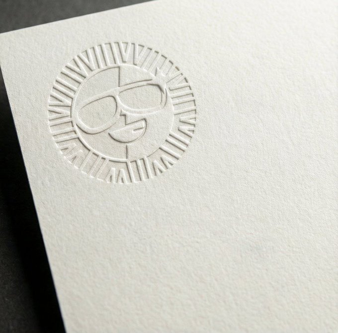 A close-up of a white textured paper card featuring a blind embossed logo of a face with sunglasses inside a sunburst design.