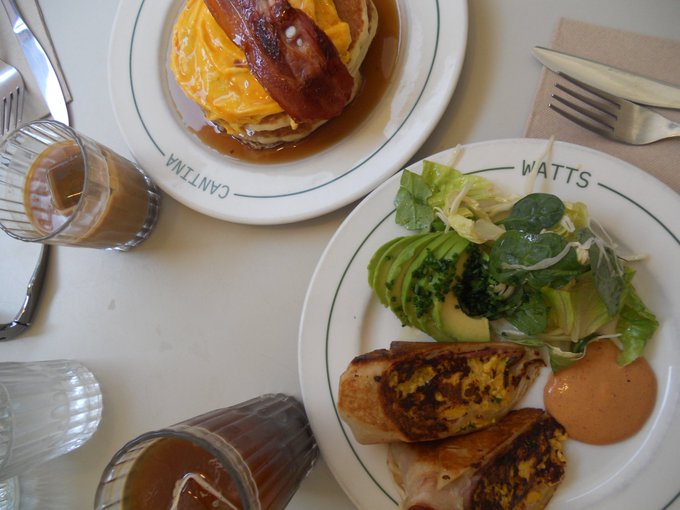 Two brunch plates at restaurant WATTS containing eggs benedict with bacon, an omelet, avocado salad, and breakfast burritos, alongside glasses of coffee and a carafe.