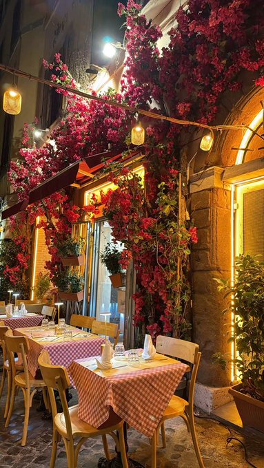 An outdoor restaurant patio at night, decorated with abundant pink bougainvillea flowers, string lights, and wooden chairs around tables with red and white checkered tablecloths.