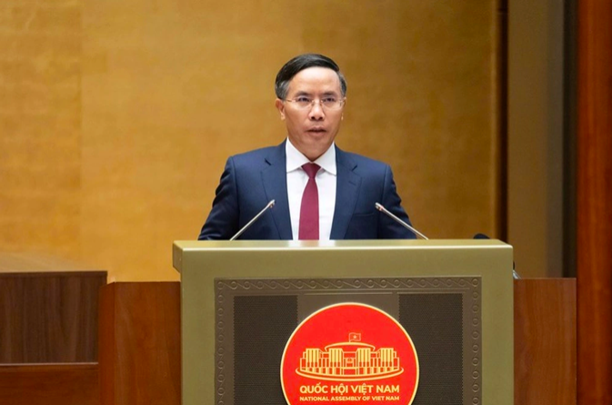 A man in a dark suit and red tie speaks at a podium bearing the emblem of the National Assembly of Vietnam, with 'QUỐC HỘI VIỆT NAM' and 'NATIONAL ASSEMBLY OF VIET NAM' written below the seal.