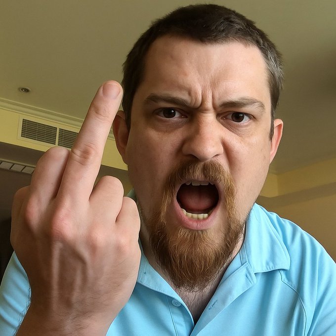 Close-up of a man with a reddish goatee in a light blue polo shirt, angrily shouting with his mouth wide open while raising his middle finger.