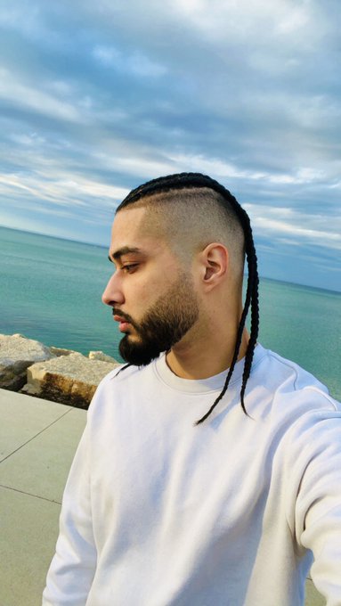 A man with braided hair and a beard takes a selfie by the ocean in a white sweatshirt, greeting followers with "Gm Friends" to start the day with a calm, coastal vibe.