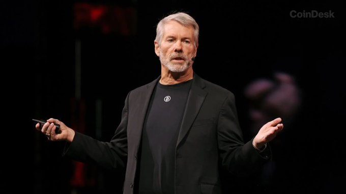 MicroStrategy founder Michael Saylor speaking on stage in a black suit jacket with a CoinDesk watermark in the corner.