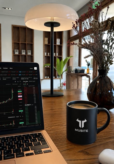 A workspace setup featuring a laptop displaying cryptocurrency price charts (including Bitcoin), a black 'MUBITE' branded mug with coffee, a modern desk lamp, and a vase of dried flowers on a wooden table.