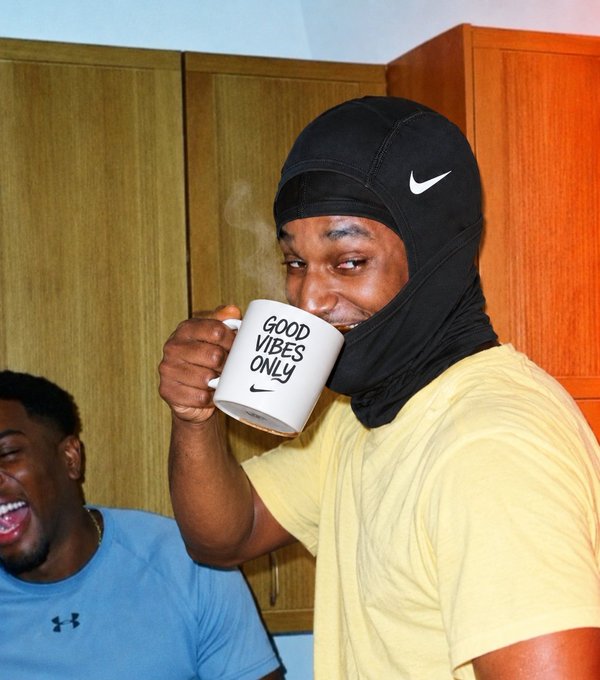 A person wearing a black Nike balaclava drinks from a white mug reading "GOOD VIBES ONLY" while another person laughs beside them.