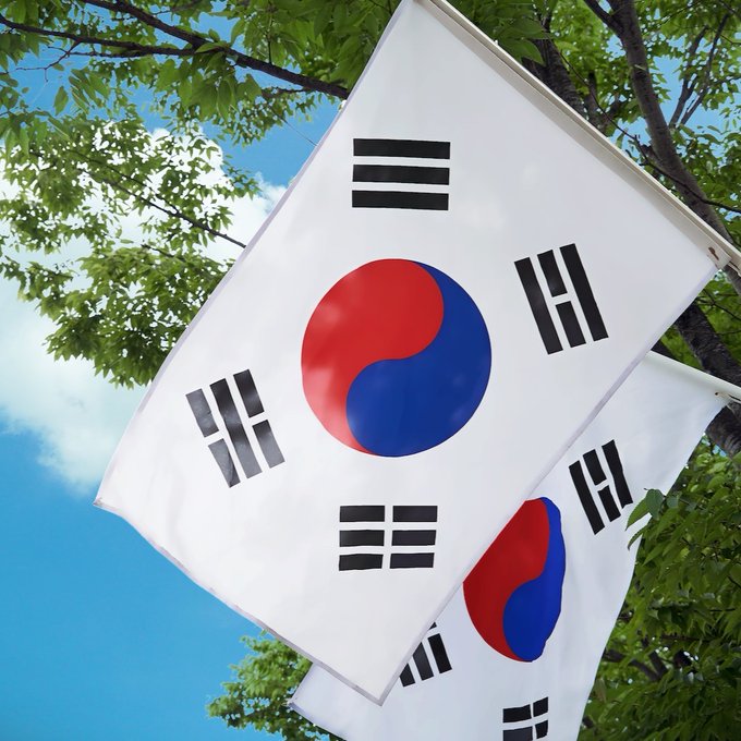Two South Korean flags wave against a blue sky framed by green tree branches. The image symbolizes South Korea, connecting to news of Shinhan Asset Management's partnership to pilot a Korean won-denominated tokenized fund.