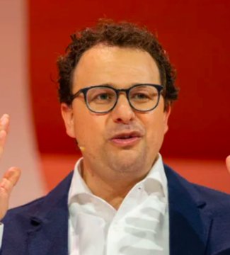 A portrait of a man with curly dark hair and glasses, wearing a white shirt and a blue blazer, speaking with his hands raised against a red background.