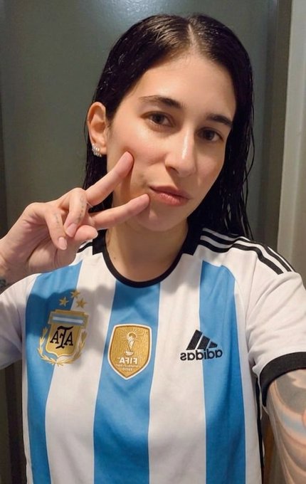 A woman with wet dark hair makes a peace sign while wearing an Argentina AFA national team soccer jersey.