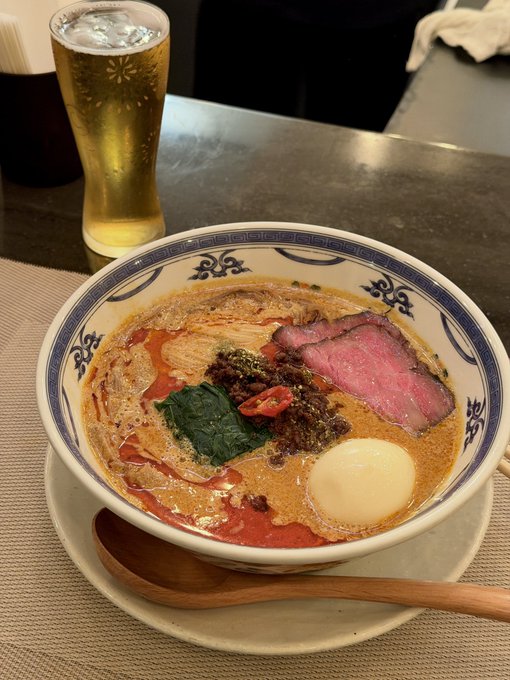 A bowl of spicy beef ramen topped with sliced beef, spinach, soft-boiled egg, and chili in a traditional blue-patterned bowl sits beside a tall glass of beer. The rich broth and hearty toppings make for a fitting "goodbye" meal to close out the #cryptosoupgroup.
