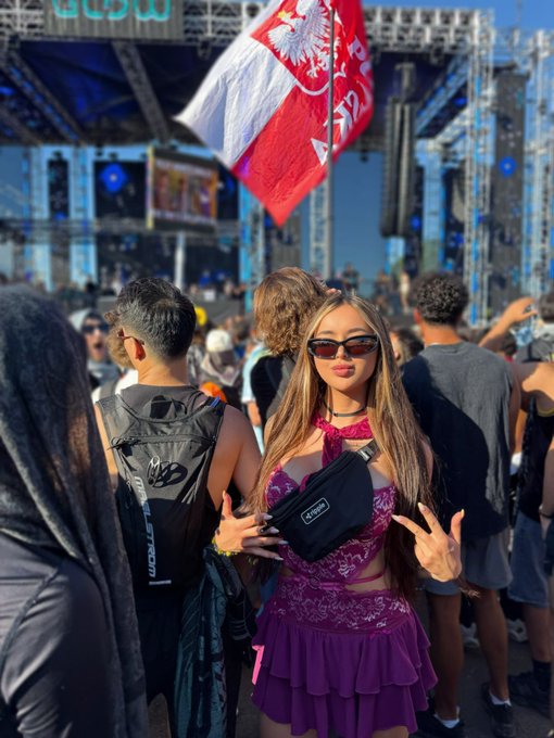 The image shows a woman wearing a purple dress, standing among several other people at an outdoor event with a large flag visible above them. The crowd appears to be enjoying themselves as they gather around the stage area. A man can be seen holding up his hand in front of him and making a peace sign gesture. "Just got back from a festival why is bitcoin down", which seems to be a reference to Bitcoin's recent drop in value, possibly indicating that she has been attending such events recently or is discussing them with others at the event.