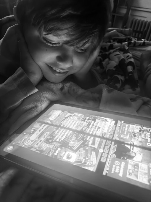 A black-and-white photograph captures a young boy lying down, smiling warmly as he looks at a tablet screen displaying a brightly lit digital interface.