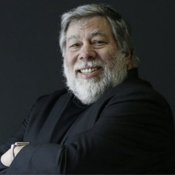 A portrait photograph of a smiling, bearded older man with gray hair, wearing a black turtleneck and dark jacket against a dark background.