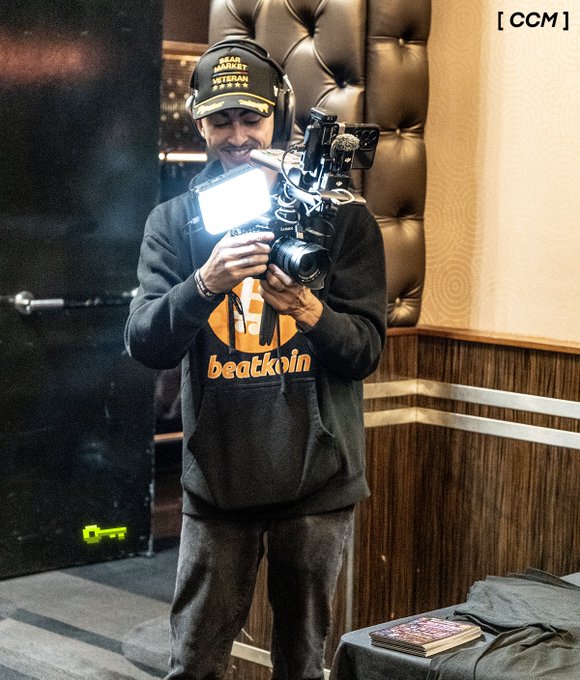 A man wearing a Sean O'Meara Veteran cap, headphones, and a black 'beatkoin' hoodie is holding a video camera with an attached light, standing in a room with a tufted leather wall and a '[ CCM ]' watermark in the top right corner.