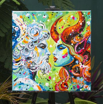 A vibrant painting on display at an easel features two stylized, colorful faces in profile—one resembling a classical bearded figure and the other a modern, flame-haired woman—surrounded by swirling patterns and rainbow hues. This artwork appears to be one of the "Angels of Freedom" pieces being showcased at the MWBTC Summit with Bitcoin For The Arts.