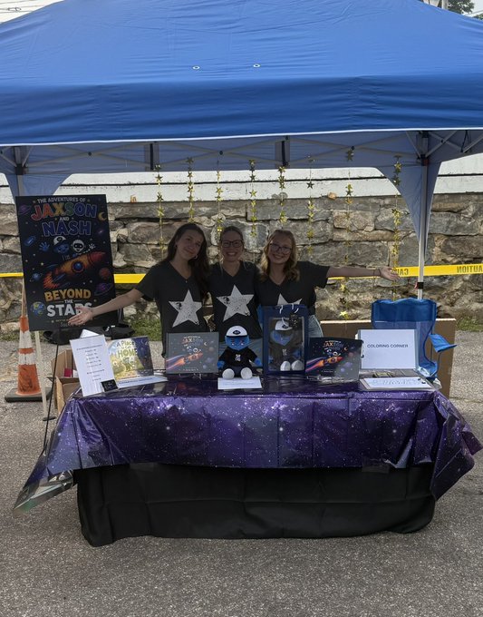 Three smiling women stand behind a vendor table draped in a galaxy-patterned cloth, displaying merchandise for "Jaxon Nash: Beyond the Stars," including a plush Space Rider toy, copies of the book, and a "Coloring Corner," capturing the joyful realization of the author whose poem has blossomed into a full creative universe.