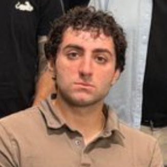 A man with curly dark hair wearing a tan polo shirt looks directly at the camera with a neutral expression.