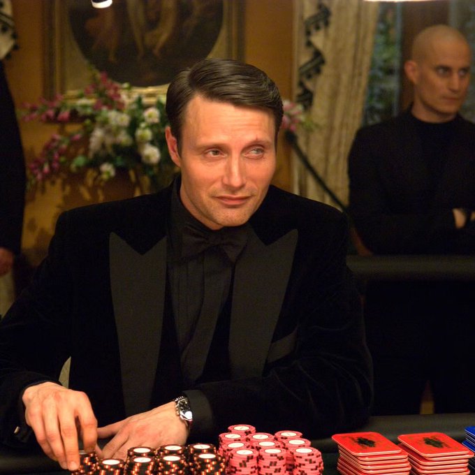 A man in a black tuxedo with a bow tie sits at a poker table, looking to the side, with stacks of red and blue chips in front of him.