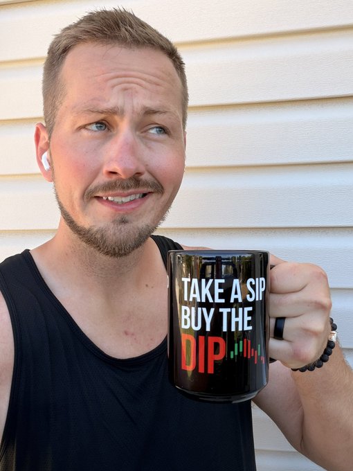 A man with a beard and a black tank top holds up a black mug with a graphic that reads 'TAKE A SIP BUY THE DIP' along with a candlestick chart pattern in red and green.