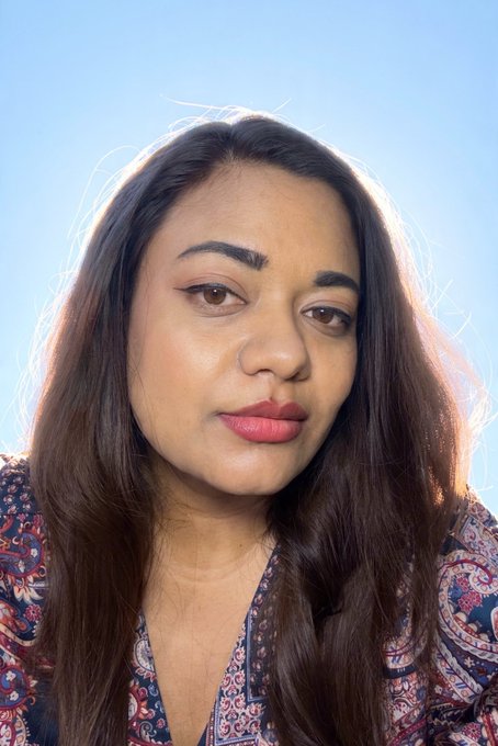 A close-up portrait of a woman with long dark hair, red lipstick, and a patterned blouse against a blue sky background.