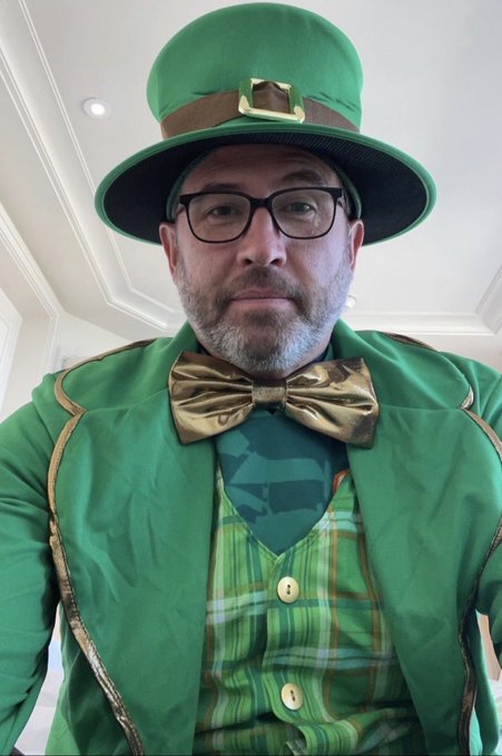 A man wearing glasses, a green top hat with a gold buckle, a green jacket with gold trim, and a metallic gold bow tie, posing for a selfie.