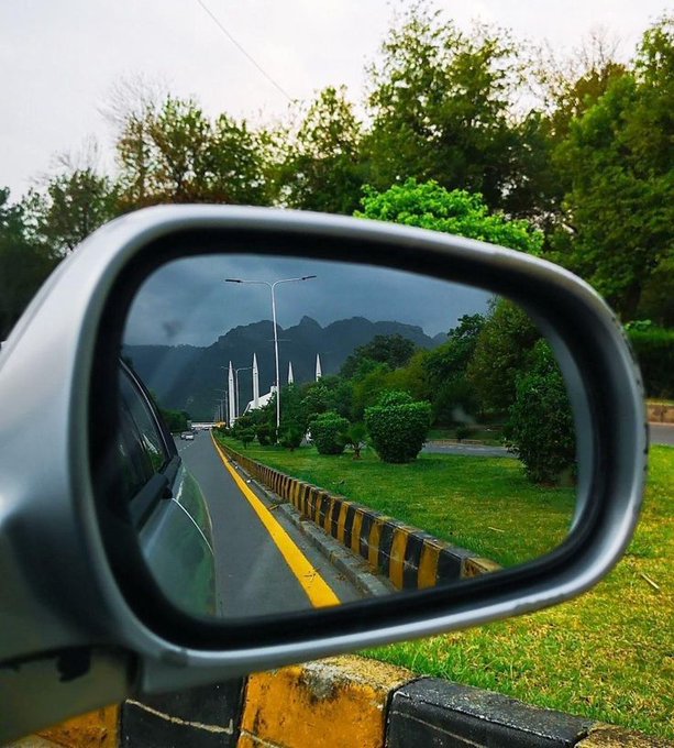 iced car windshield with a reflection of trees and mountains, viewed from the passenger side mirror on a road lined with yellow curb and green grass.